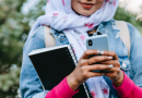 Young woman using phone with leafy background, holding a notebook and wearing a headscarf and denim jacket