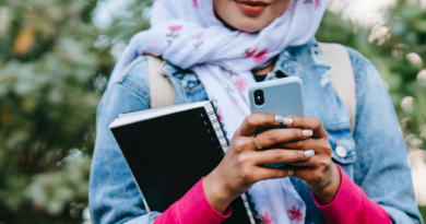 Young woman using phone with leafy background, holding a notebook and wearing a headscarf and denim jacket