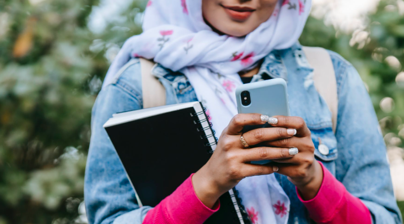 Young woman using phone with leafy background, holding a notebook and wearing a headscarf and denim jacket