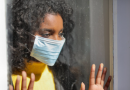 Young woman wearing mask looking out of window longingly with hands pressed against it