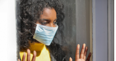 Young woman wearing mask looking out of window longingly with hands pressed against it