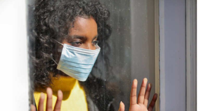 Young woman wearing mask looking out of window longingly with hands pressed against it