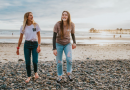 Two young women walking along a pebbled beach laughing