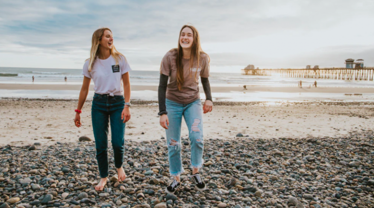 Two young women walking along a pebbled beach laughing