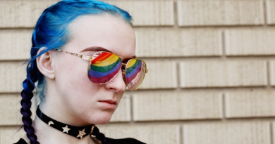 An 18 year old young person with blue hair in plaits wearing sunglasses with the pride flag reflecting in their sunglasses