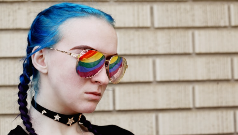 An 18 year old young person with blue hair in plaits wearing sunglasses with the pride flag reflecting in their sunglasses