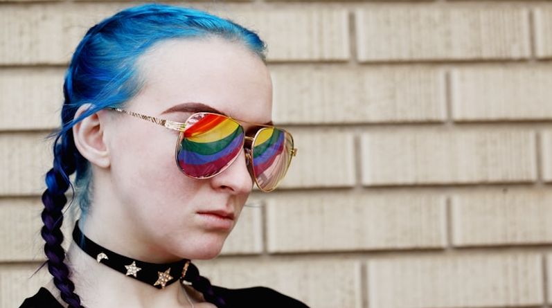 An 18 year old young person with blue hair in plaits wearing sunglasses with the pride flag reflecting in their sunglasses