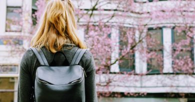 Young woman with backpack facing a building with windows and a tree with pink blossom.