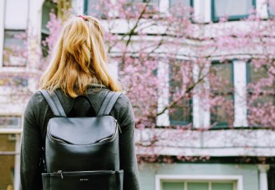 Young woman with backpack facing a building with windows and a tree with pink blossom.