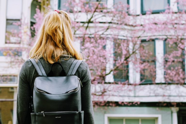 Young woman with backpack facing a building with windows and a tree with pink blossom.